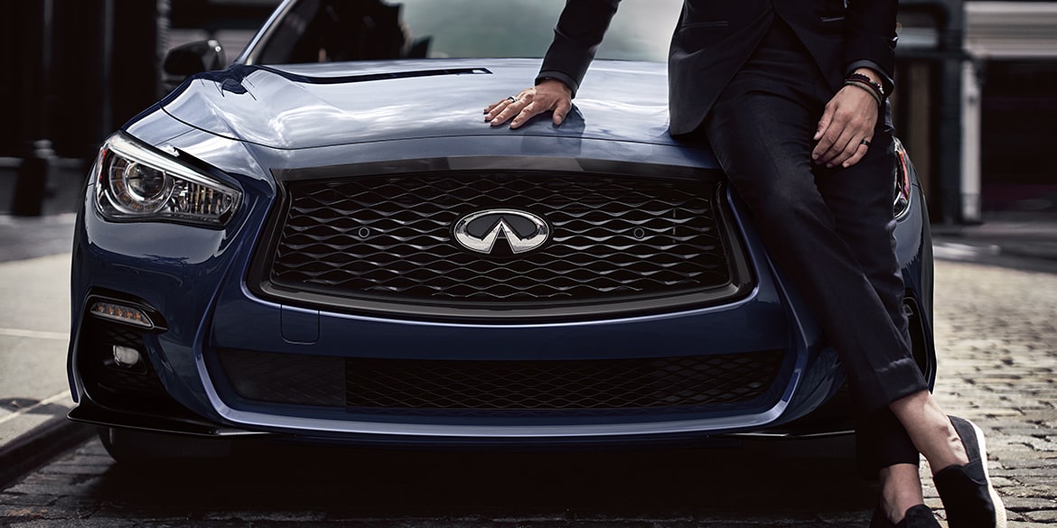 Front exterior profile view of man leaning on the hood of a 2024 INFINITI Q50 highlighting Q50 grille and LED headlights