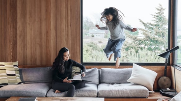 Interior view of home with woman sitting and reading book on couch while girl jumps on couch